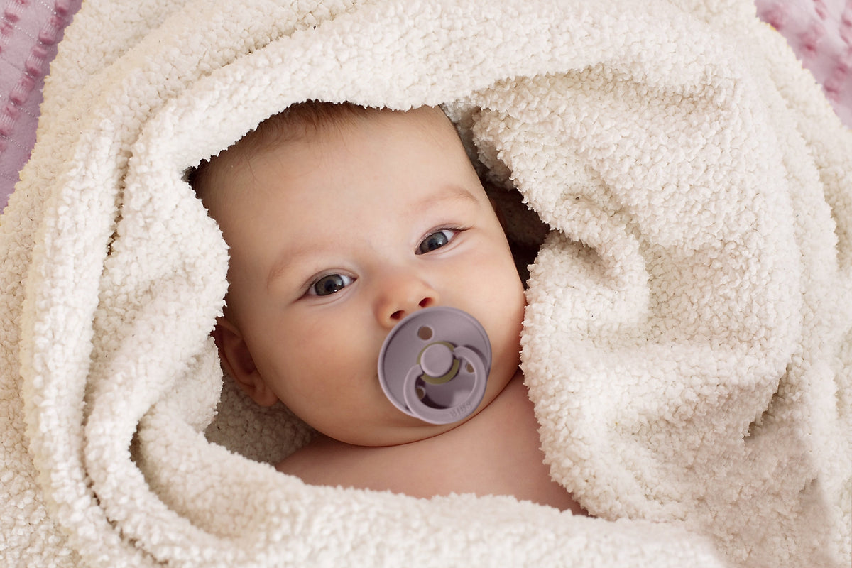 Baby met een BIBS Colour speen, gewikkeld in een zachte crèmekleurige deken.